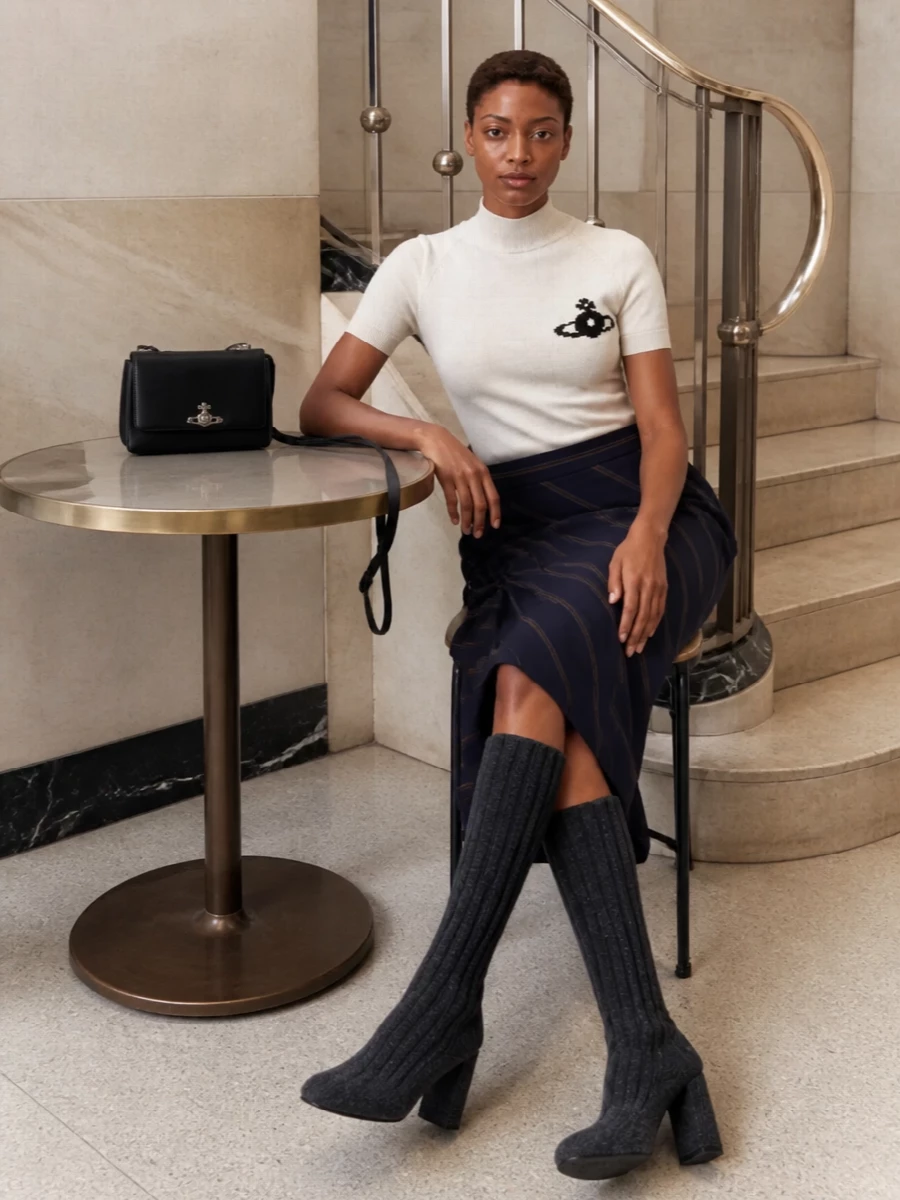 Soft ivory and inky pinstripe meet in a quietly subversive pairing that feels as at home in a gallery as a late client drink. The close-fitting high neck and sculpted draped skirt frame the waist, while charcoal knit boots and a small black shoulder bag ground the palette in precise, modern lines. It is pared back but unmistakably Vivienne Westwood, more about proportion and texture than noise.