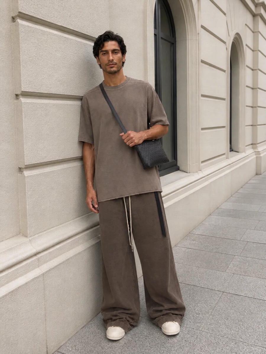 Taupe on taupe sets up a calm, underground uniform, with the longline tee extending the line of the ultra-wide trouser for a pooled, columnar profile. Cream DRKSHDW high-tops just peek out from under the hem, lightening the base while the dark woven crossbody adds a compact, utilitarian accent at the hip. The result is a minimalist Rick-centric streetwear set that feels grounded, easy and quietly directional for warm-weather city days.