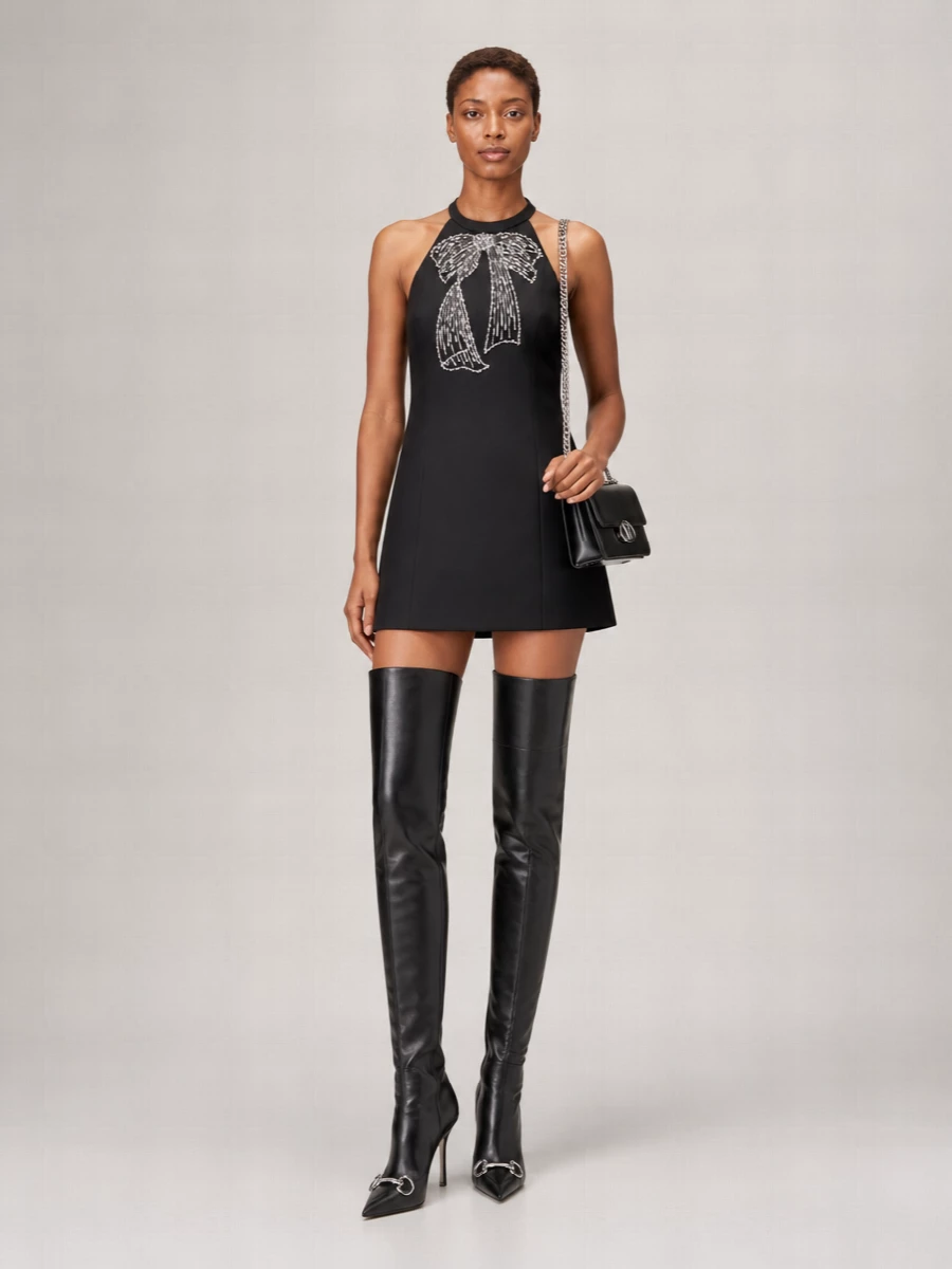This is a sharp black-on-black evening ensemble that lets the Gucci over-the-knee boots and Valentino halter mini work together as one long, graphic line of gloss and matte. The bare leg between hem and boot shaft keeps it sensual but contained, while the small structured shoulder bag adds just enough silver hardware to echo the boot detail without tipping into ornament. Choosing the dress path over tailoring preserves the simplicity and impact of the silhouette, keeping the mood modern, precise and unmistakably night-focused.