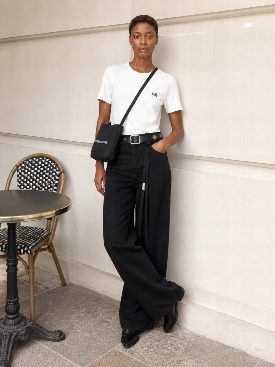 A soft white Ann Demeulemeester tee meets high-waisted black wide-leg trousers for a monochrome line that feels calm, graphic and quietly offbeat. Pointed black loafers and a slim studded belt sharpen the proportions, so the volume reads intentional rather than relaxed. A simple black cross-body finishes the outfit with just enough logo to keep it recognisably directional, but still low-key for everyday city wear.