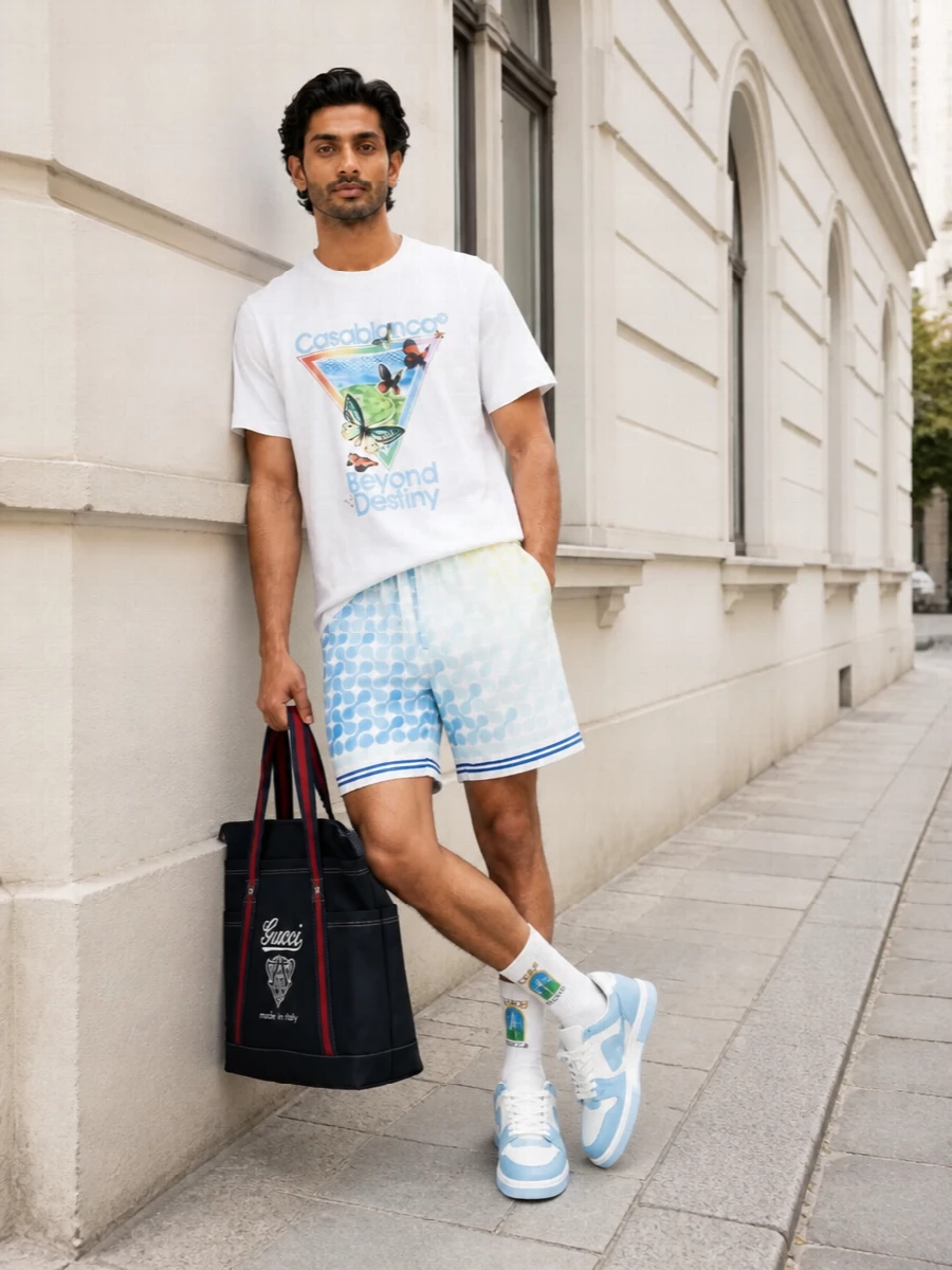 A sun-drenched graphic tee sits over soft-geometry shorts in washed sky and lemon, so the palette feels light, playful and vacation ready. Powder-blue court sneakers and a navy Gucci tote keep the energy in that tennis-resort sweet spot, sporty but still polished. Logo socks peeking above the sneakers are the only extra detail, keeping the whole outfit clean despite the color story.