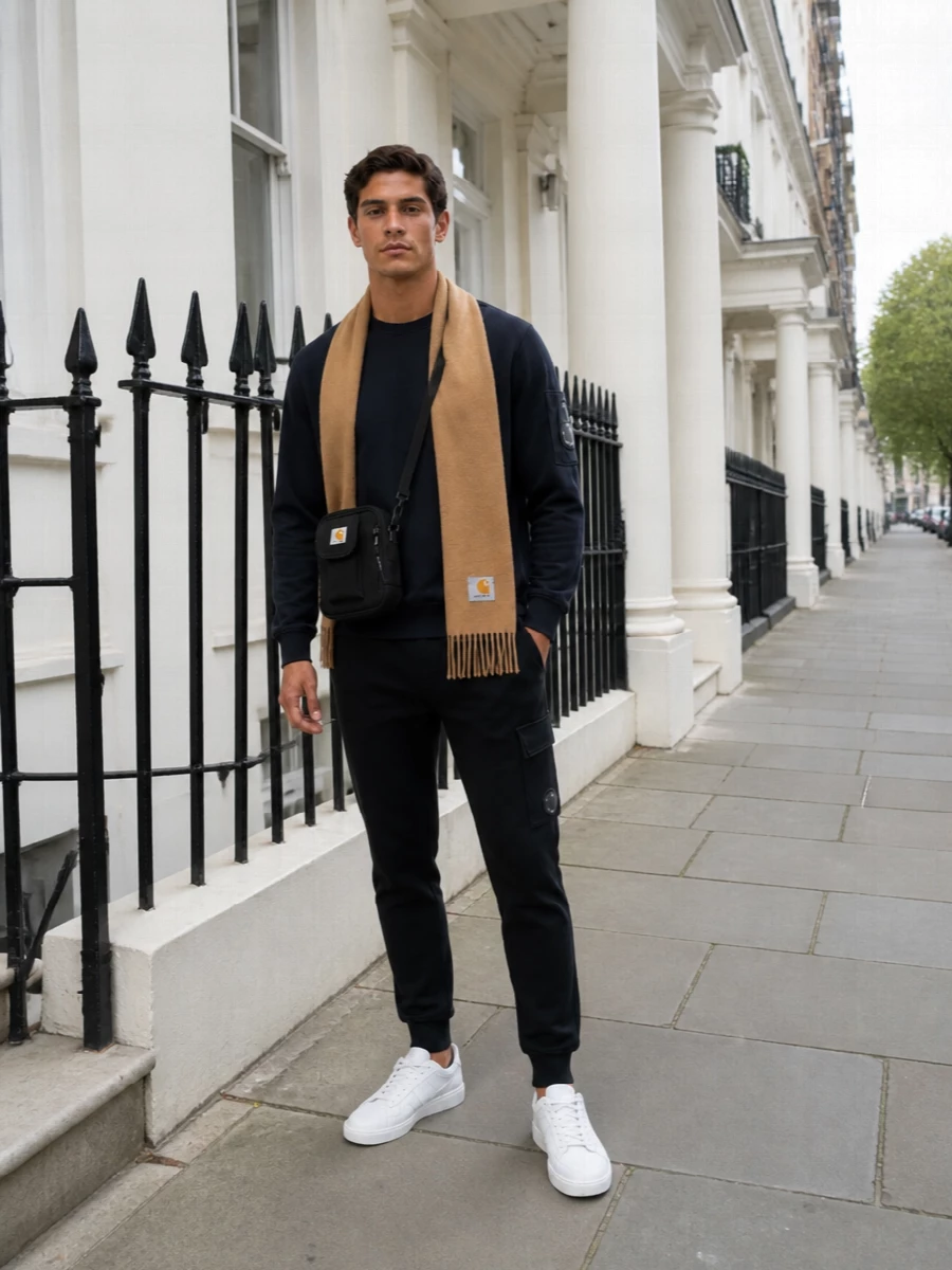A deep navy C.P. Company crew with sleeve lens runs straight into matching black lens-detail joggers for a clean, branded trackset feel that stays low-key. Crisp white Comme des Garçons sneakers sharpen the base and keep it athletic, while the compact black Carhartt crossbody and camel scarf add just enough city utility and warmth without diluting the technical mood.