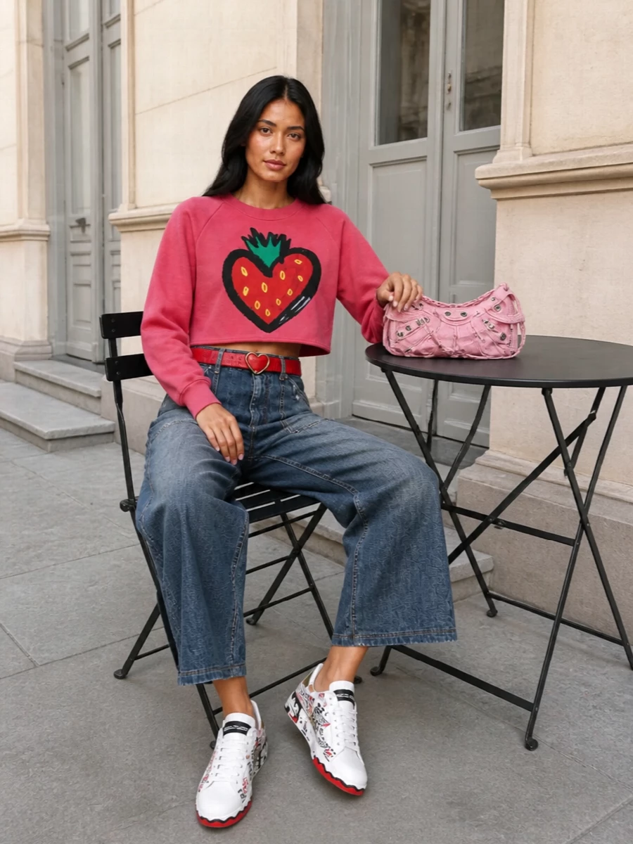 A cropped strawberry sweatshirt sits over sculptural high-rise wide-leg denim, creating a punchy proportion that feels playful yet grounded. Heart-detailed sneakers and a bubblegum shoulder bag keep the story fun and graphic. The lipstick-red heart belt nods to the print and pulls everything into a cohesive Dolce romance-meets-streetwear moment.