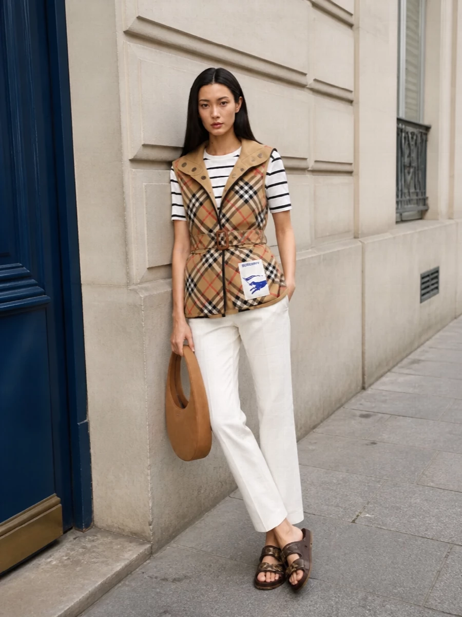 I’m styling the Burberry sleeveless trench as the hero over a streamlined top-and-trouser base rather than a dress, to keep the look sharp, versatile and true to its preppy–utility energy. The striped Max Mara knit and ivory tailored trouser create a clean, elongating column under the short, belted layer, while Burberry’s rich brown slides and a camel Coperni bag quietly echo the heritage tones. The result is an SS city uniform: graphic, polished and relaxed enough for all-day wear without losing that luxury bite.