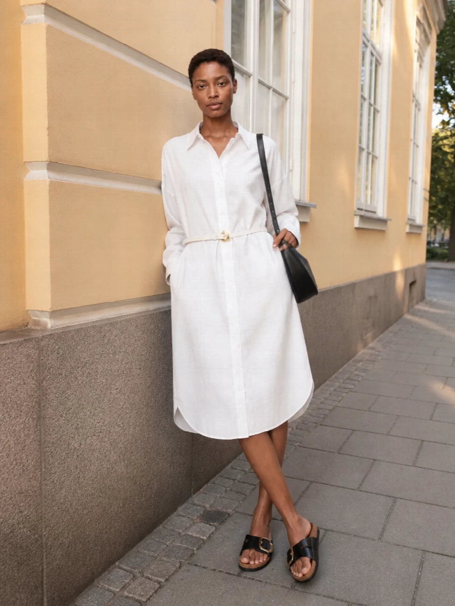 A crisp white shirt dress keeps the focus on line and proportion, letting the black cork slides ground everything with subtle hardware detail. The curved black shoulder bag ties back to the sandals, giving a restrained black-and-white palette that feels quietly assured from market mornings to terrace dinners. Choosing the dress route keeps the outfit minimal and unified, which suits the easy, flat sandals better than breaking the body with a separate top and bottom.