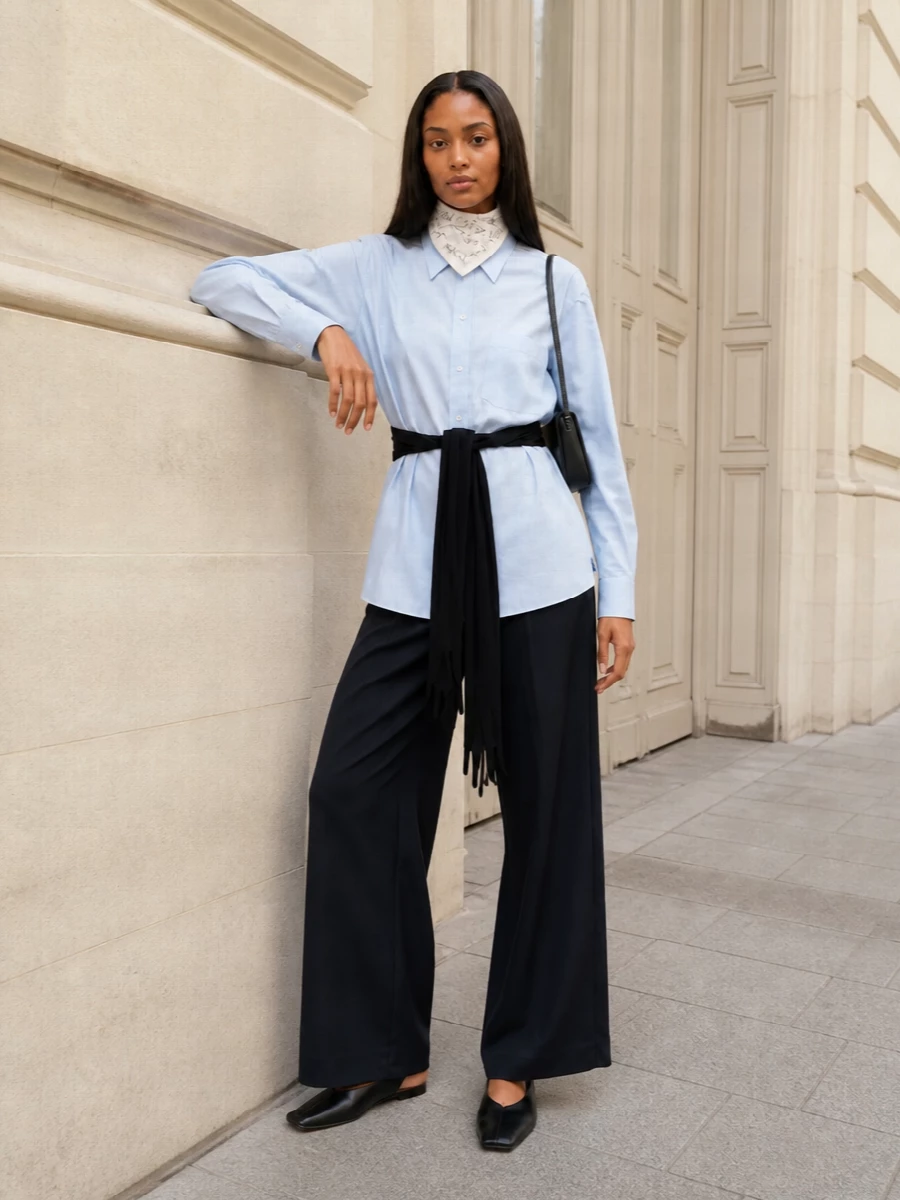 Pale blue and deep navy set up a calm, almost academic palette, with the glove-tied shirt cinched over fluid wide-leg tailoring for a long, precise line. Black slingback flats and a slim shoulder bag keep everything pared back and functional, letting the subtle subversion of the waist detailing stay in focus. The silk scarf is a final, quiet nod to Margiela’s cerebral side, whether at the neck or knotted to the bag.
