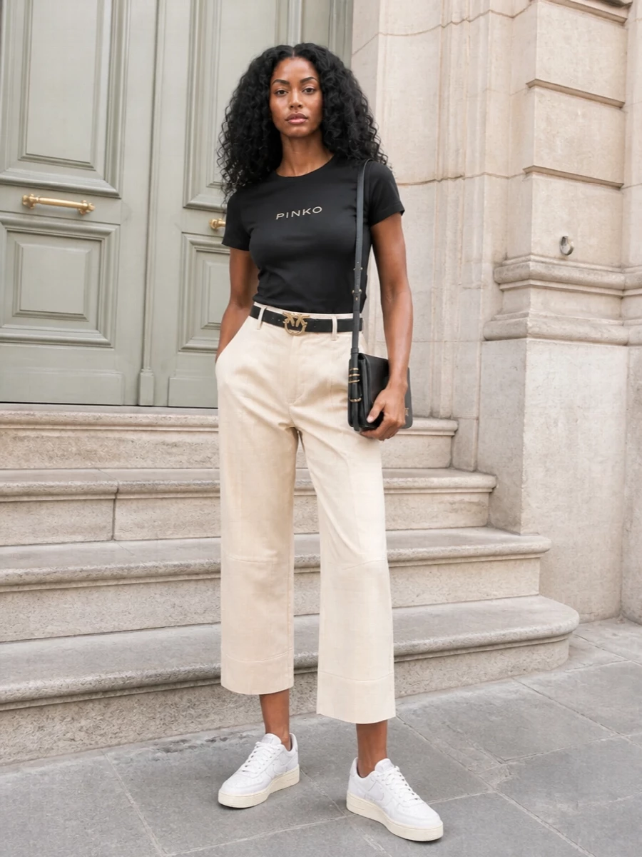 A black logo tee meets soft beige tailoring and creamy low-top sneakers for a clean, city-ready mix of streetwear and preppy ease. The black shoulder bag and bird-buckle belt quietly echo the tee’s branding, pulling the palette together without shouting. It is the kind of outfit that works for relaxed office hours, weekend plans, or any day you want to look put-together but still feel off-duty comfortable.