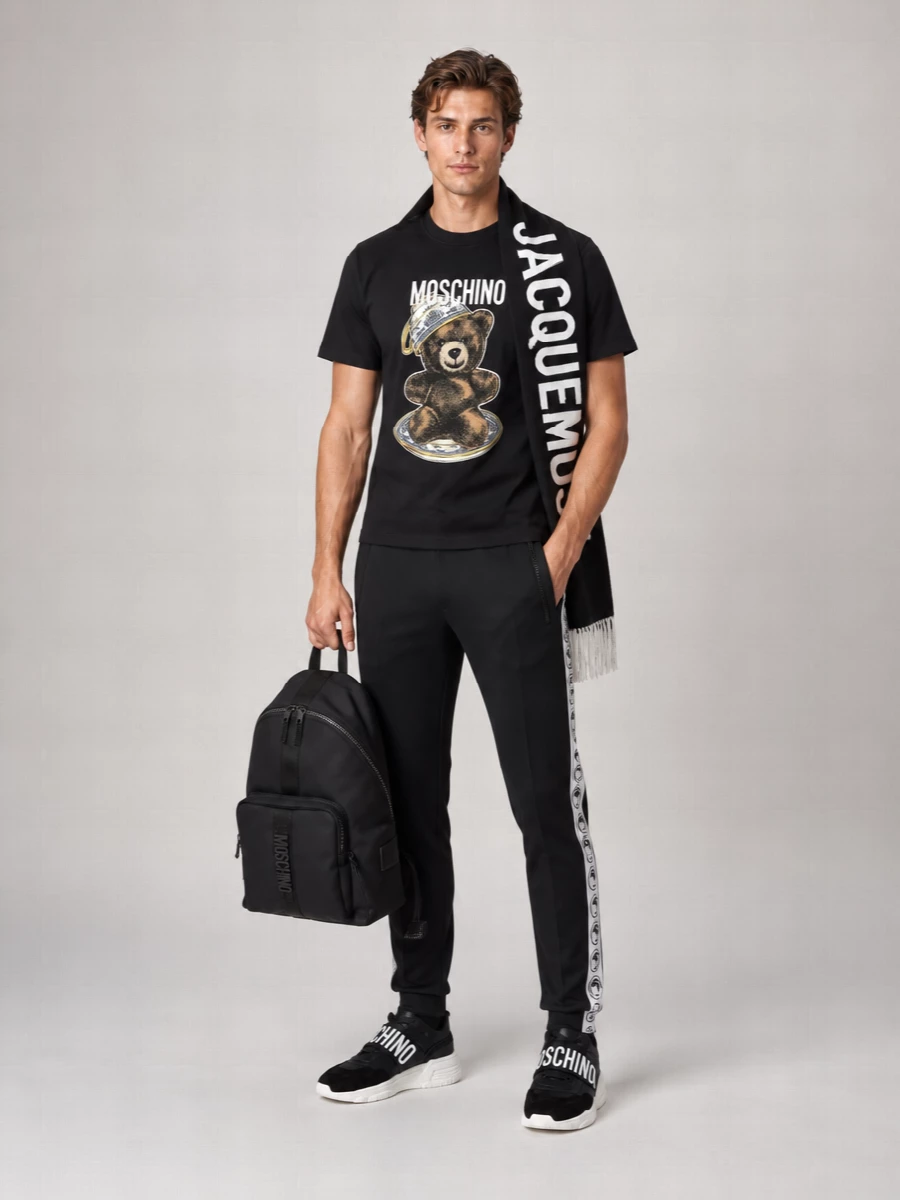 Moschino’s teddy tee stays front and center, framed by sharp black logo-stripe track pants for a clean, athletic base. Black low-top sneakers and a tonal backpack keep the story streamlined, while a bold black and white logo scarf adds winter function and a final graphic hit without crowding the print.
