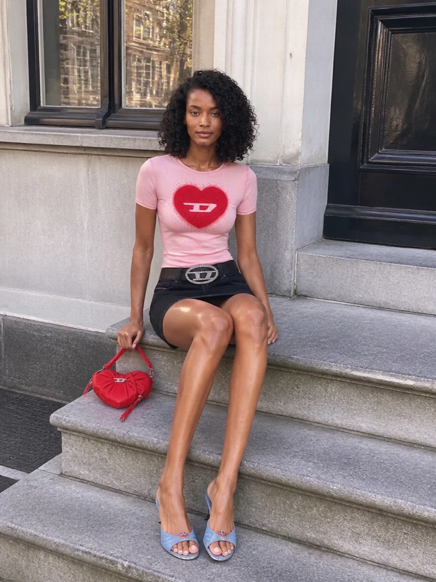 A curve-hugging pink logo tee tops a washed black low-rise mini for that early‑2000s club energy, all legs and attitude. Light denim kitten mules keep the palette soft and nostalgic while still ready for a dance floor. A crystal‑buckled belt and glossy red heart bag double down on the playful Diesel branding and tie the red accents together without feeling overdone.