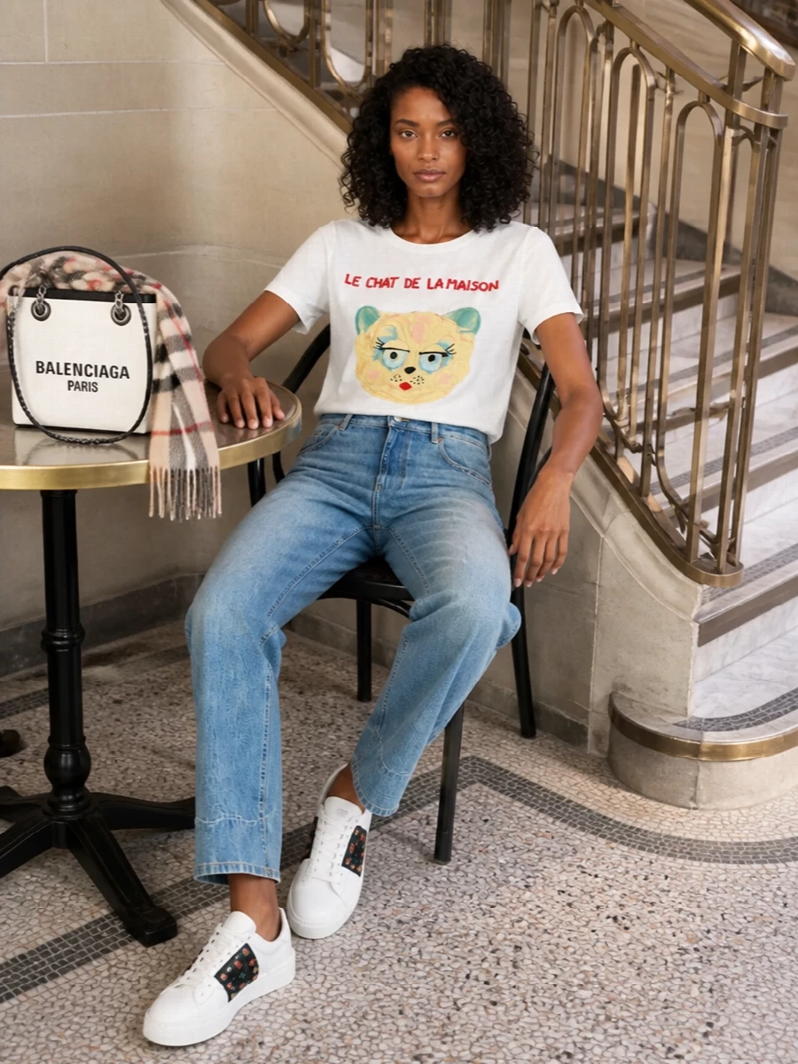The Valentino cat tee sits atop precise, softly faded straight-leg jeans for a relaxed but considered base that keeps the focus on the playful graphic. Cherry-accented white sneakers and a crisp off-white mini tote echo the tee’s palette, while a gauzy Burberry check scarf adds a final preppy note that feels effortless for FW city days. The result is a polished streetwear look with a witty, Parisian twist.