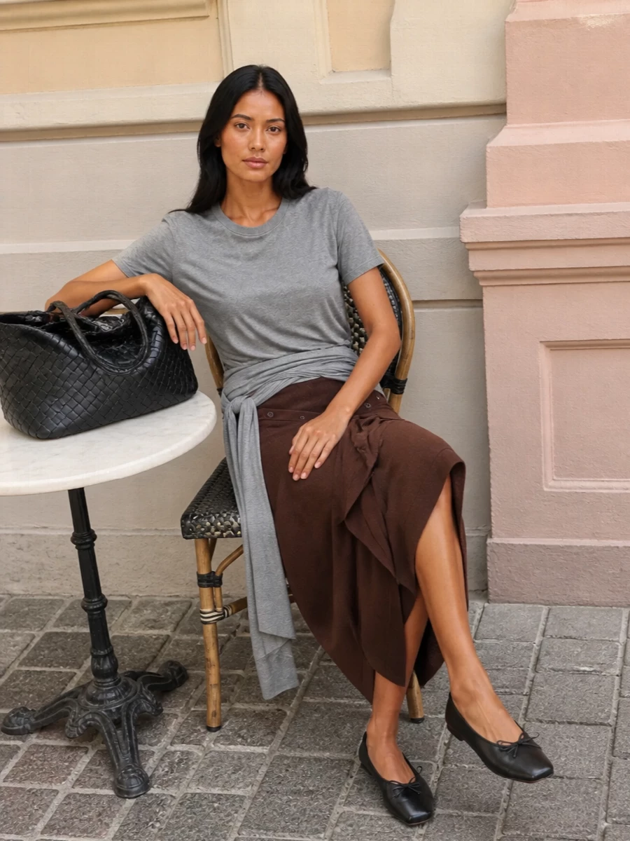 Heather grey jersey and chocolate knit meet in a fluid, sculpted pairing that feels relaxed but quietly intentional. The asymmetric tee and wrap skirt play off each other’s drape, creating soft movement rather than strict tailoring. Black, minimalist accessories sharpen the mood just enough while the woven tote brings in a tactile, boho touch that keeps the outfit grounded and easy for city days.