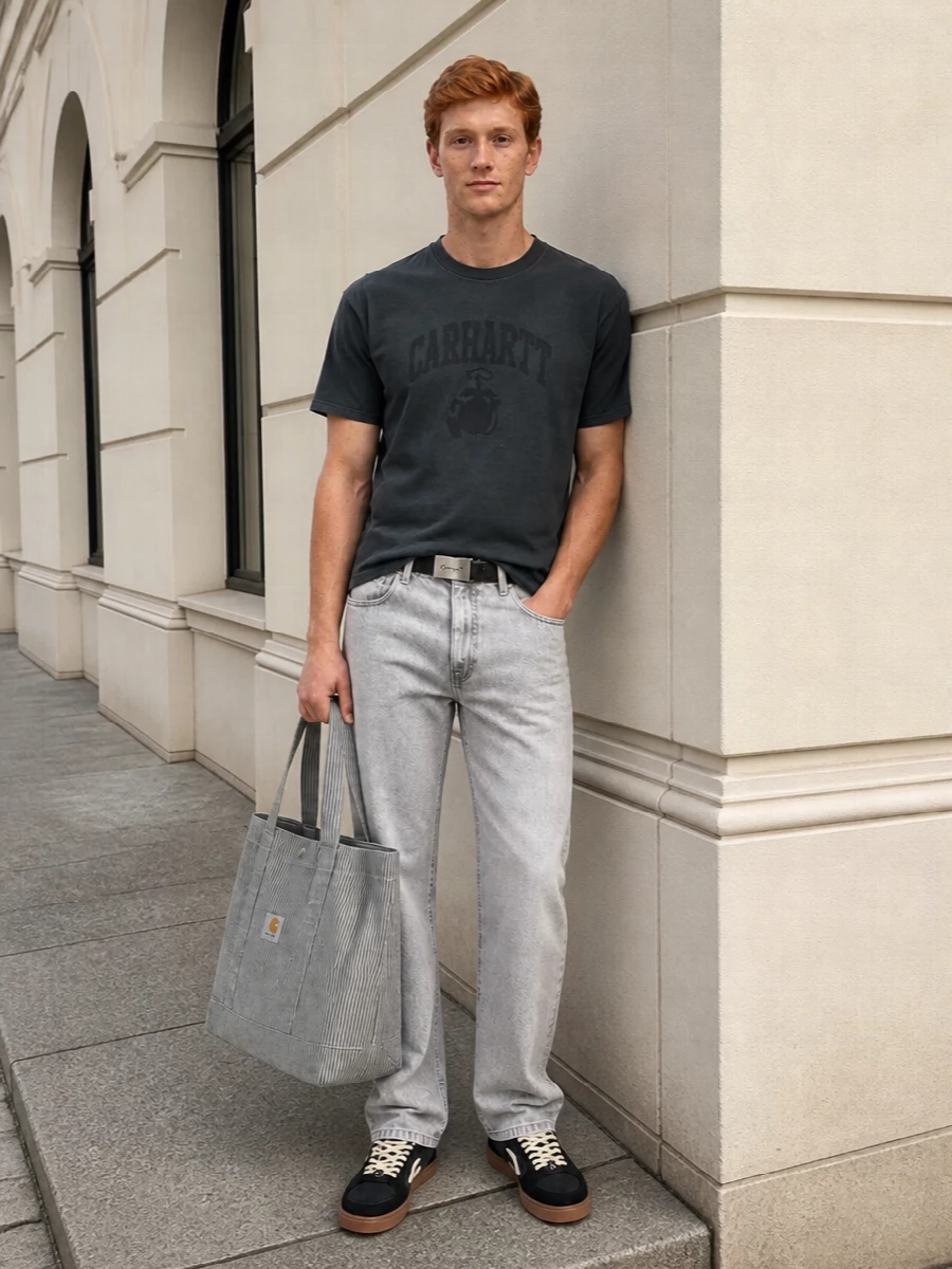 A washed charcoal tee sits against light grey straight-leg denim, keeping the palette muted while adding just enough contrast to feel considered. Off-White skate sneakers pull the dark tones downward, with cream and gum hits softening the workwear edge. A striped Carhartt tote and a slim Margiela belt finish it with graphic practicality and a small injection of quiet designer detail.
