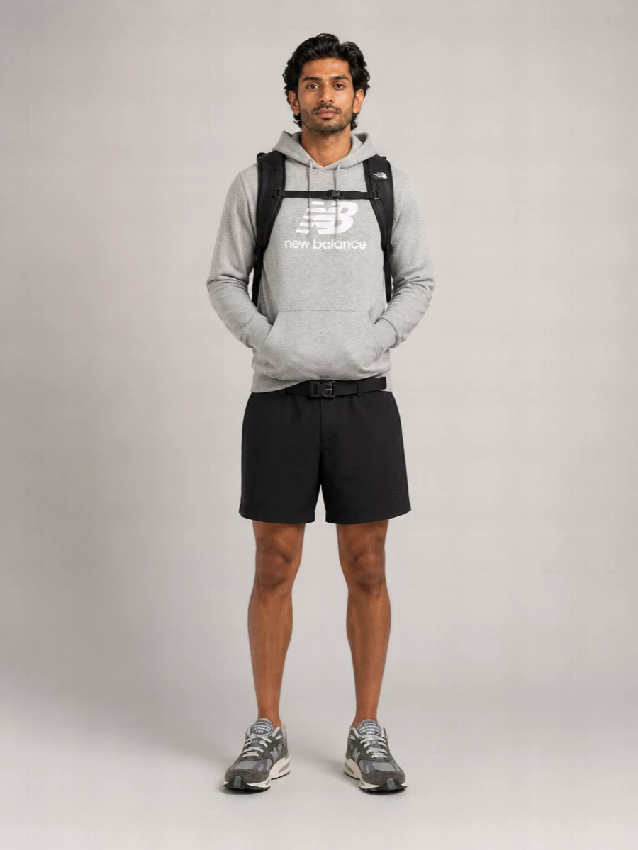 A heather grey New Balance hoodie sits against clean black shorts for a sharp, minimal take on athleisure. Tonal grey New Balance runners keep the line sleek and low-key, while a technical black backpack reinforces the sport-utility attitude. The elastic utility belt gives a discreet hit of polish without disturbing the relaxed, off-duty feel.