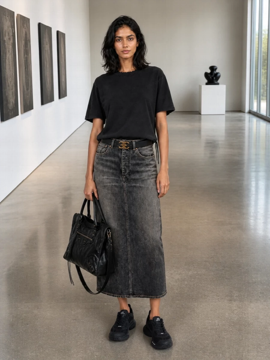 A washed-black lace-up tee meets a slim, vintage-faded denim midi for a long, grunge-leaning silhouette that still feels precise. Chunky black runners and a lived-in leather tote keep it firmly in the city, relaxed enough for day but strong enough for late nights. The discreet logo belt adds just enough structure at the waist without breaking the monochrome line.