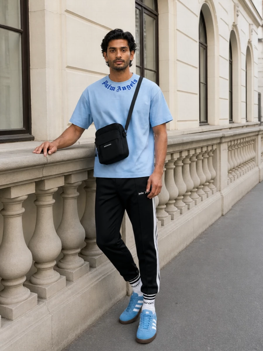 Pale sky blue up top gets anchored with sharp black track pants for a clean, sport-driven street mix. Sky suede sneakers and striped socks echo the tee’s blue tones, keeping everything light but intentional. A compact black crossbody finishes it with just enough logo attitude for city days or late-night runs.