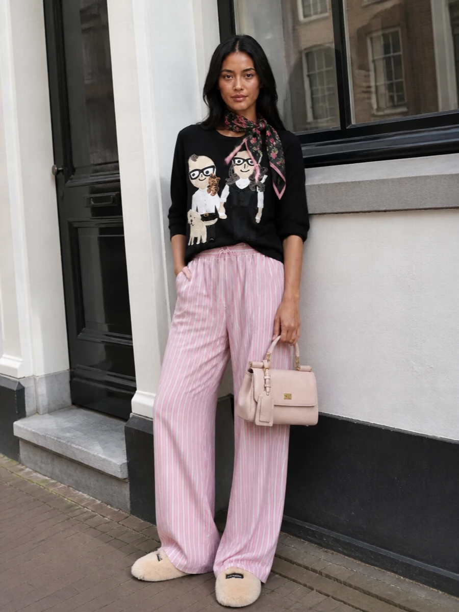 A storybook lounge tee meets soft pink striped trousers for a romantic, off-duty set that feels playful yet composed. Cream faux-fur slides and a blush top-handle bag keep the palette gentle, while a rose-print scarf subtly links the black and pink tones for a quietly cohesive finish. Ideal for days that drift between home and casual errands without changing out of something you love.