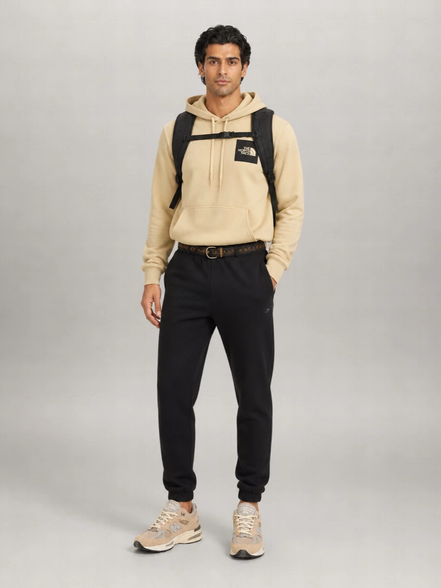 A sand hoodie sits against deep black joggers for a simple, graphic base that feels clean rather than lazy. Neutral, cushioned runners in soft taupe keep the line athletic and modern while echoing the hoodie’s warmth. A structured black backpack and slim webbing belt sharpen the mood just enough for city days and travel.