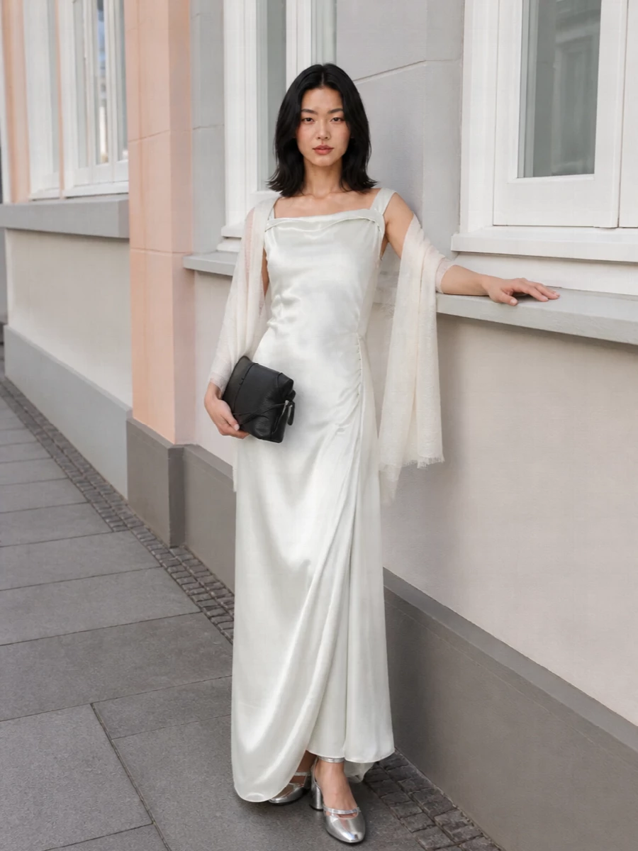 An ice-pearl column of satin is grounded with a slim black leather clutch, keeping the palette precise and the mood quietly formal. Silver block-heel straps catch the light as you move, echoing the gown’s liquid sheen. A gauzy ivory stole skims bare shoulders, adding just enough softness for a gallery opening or black-tie dinner without cluttering the line.
