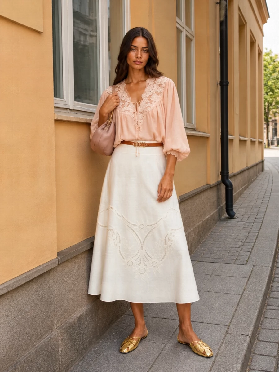 A blush lace blouse floats over a softly structured ivory embroidered midi, creating a romantic silhouette that feels light yet considered for warm-season evenings. Gold cut-out flats and a ruched blush clutch bring a subtle glow without tipping into formality, while a slim tan logo belt quietly defines the waist. The result is feminine and bohemian, but polished enough for garden parties, dinners or gallery nights.