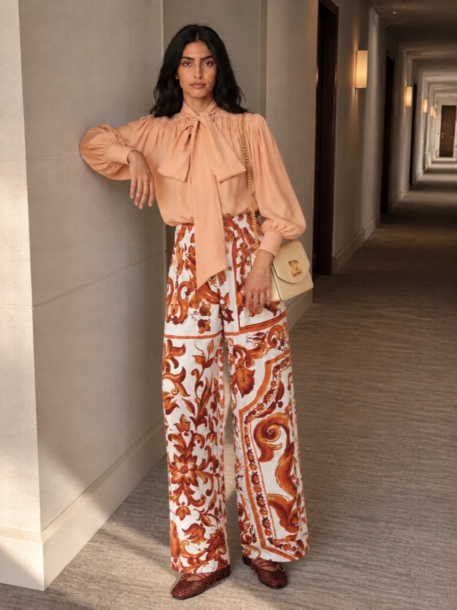 Romantic but grounded, this pairing lets the baroque terracotta print stay center stage against a whisper-light peach bow blouse and barely-there mesh ballerinas. The warm buttercream shoulder bag ties into the soft neutrals, while gold hardware and delicate straps add just enough formality for intimate dinners or gallery evenings. Everything moves and drapes, so the wide leg reads languid rather than loud.