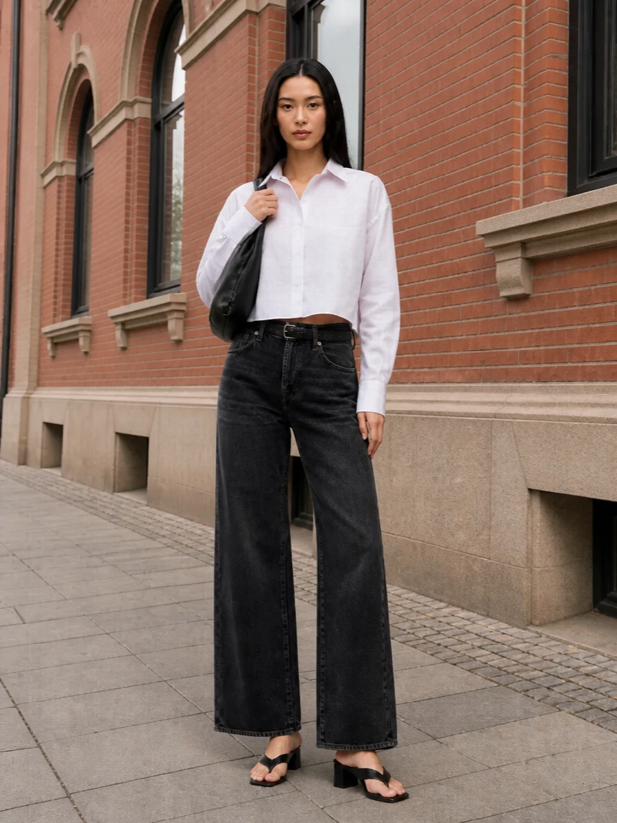 Washed black wide-leg denim meets a crisp cropped white shirt for a clean, modern base that feels sharp without being fussy. Square-toe black mules and a sculptural black shoulder bag keep everything grounded in a monochrome, Margiela-leaning palette. A slim black belt just snips in the waist so the volume reads intentional, not oversized by accident.