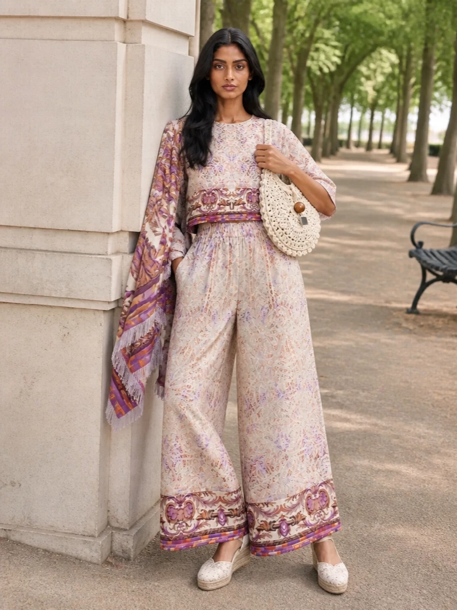 A romantic, bohemian co-ord in soft ecru and orchid creates a fluid column, the cropped blouse neatly framing the waist above those whisper-light wide legs. Neutral lace espadrille wedges and a sculptural crochet bag keep the focus on print and movement while adding tactile interest. A baroque-inflected wrap in matching purples and golds finishes the story with a gentle, artful flourish for sunlit days and warm evenings alike.