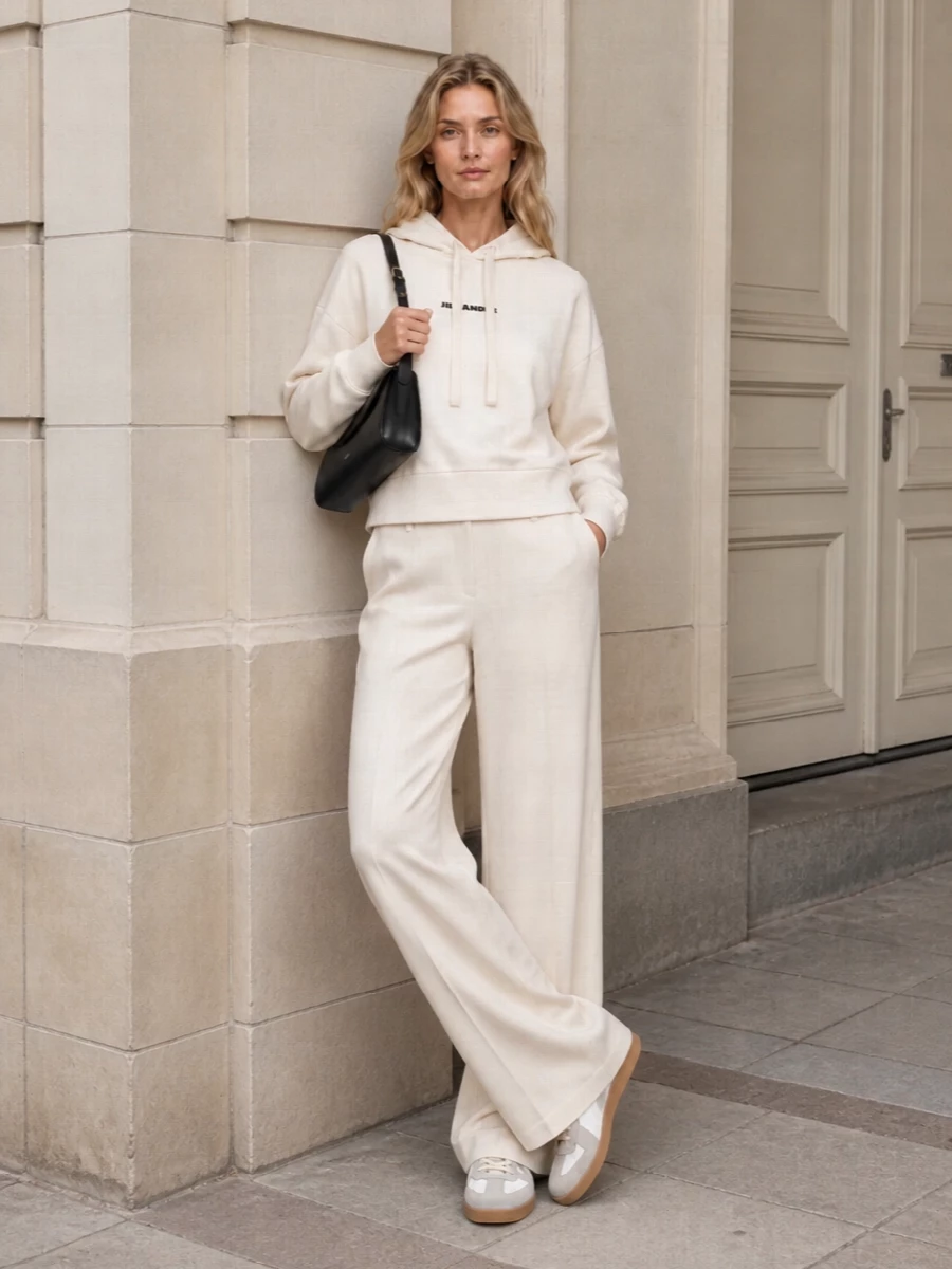 A cream Jil Sander hoodie meets fluid ivory tailoring for a soft, elongated column of neutral color that feels calm and quietly precise. Margiela’s pared-back sneaker keeps the mood relaxed yet intelligent, while the structured black Jil Sander shoulder bag adds a single graphic line to anchor all that ease. It is a minimalist city uniform that reads considered without ever trying too hard.