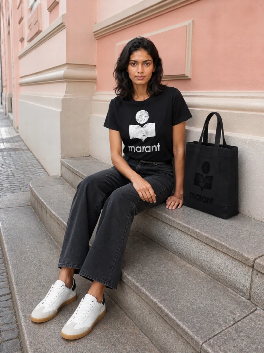 Ink logo, washed charcoal denim and crisp white sneakers keep the outfit graphic but easy for everyday city wear. The straight-leg jean and slim black belt give just enough structure to balance the relaxed tee. A logo tote finishes it with practical scale and a discreet nod to the brand.
