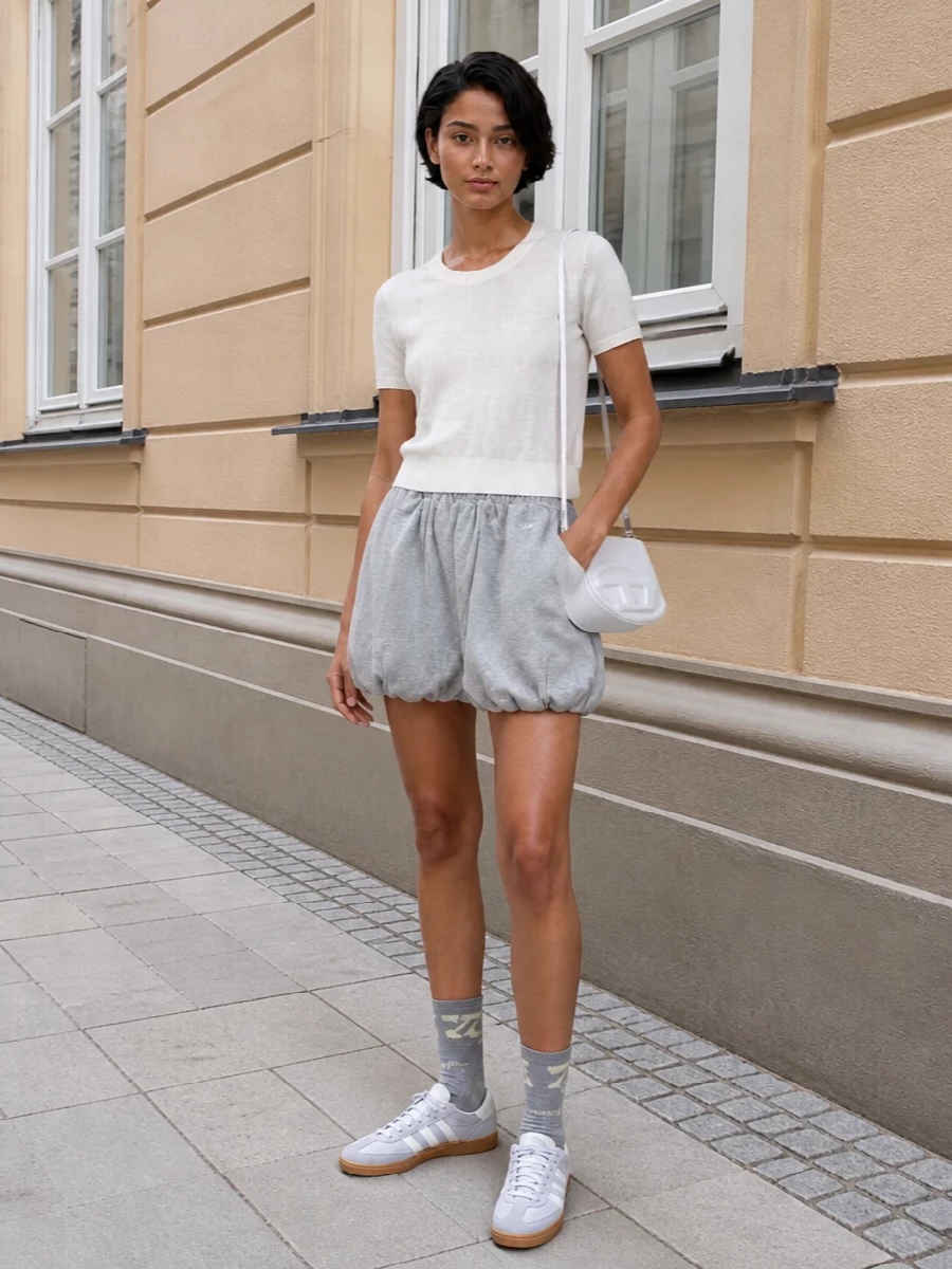 A soft white knit tee and heather grey bubble shorts build a clean, sport-influenced base in cool neutrals. Slim retro sneakers and a structured white cross-body keep everything light, sharp and easy for off-duty days. Grey logo socks peek out just enough to connect the palette and add a quiet street detail.