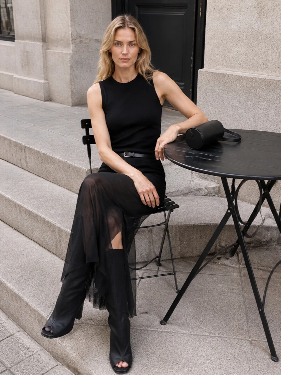 A sharply sculpted black column anchors this evening ensemble, all clean lines above and gossamer movement below. Sculptural wedge boots and a narrow belt emphasize the vertical, architectural silhouette, while a discreet cylindrical shoulder bag keeps everything focused and quiet. The result is dark, intelligent glamour that feels deliberate rather than over-styled.