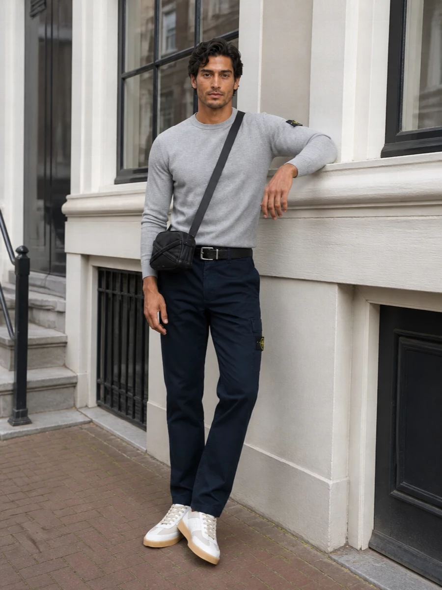 Navy Stone Island cargos are cleaned up with a soft grey knit, keeping the line relaxed but intentional. Taupe and white gum-sole sneakers lighten the palette, while a compact black crossbody and precise belt give the city-ready finish. The result is easy technical streetwear that still feels considered enough for all-day movement between errands, commute and casual plans.