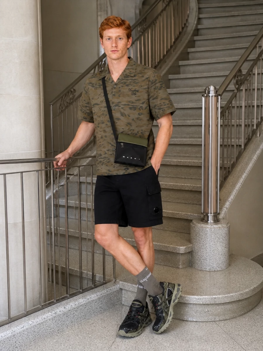 Carhartt WIP’s diffuse camo shirt stays in focus against clean black C.P. Company shorts, keeping the street-utility energy sharp but unfussy. Taupe and black Asics runners and matching Marni crossbody echo the earthy palette with a technical twist, while logoed socks add a low-key brand hit that finishes the frame. The result is an easy, city-ready uniform that moves from skate spot to late bar without a change-up.