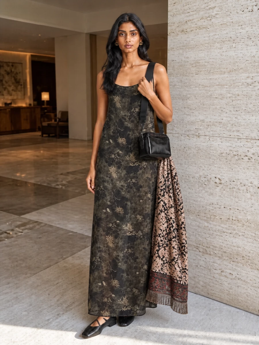 A shadowy floral column pairs with sculptural black flats and a glossy black shoulder bag for a grounded, poetic take on day-to-dinner dressing. Gold studs bring just enough light to the face, while a muted floral scarf can be draped or tucked away to shift the mood from relaxed day to more focused evening. The result feels moody, romantic and quietly precise around the long, diaphanous silhouette.