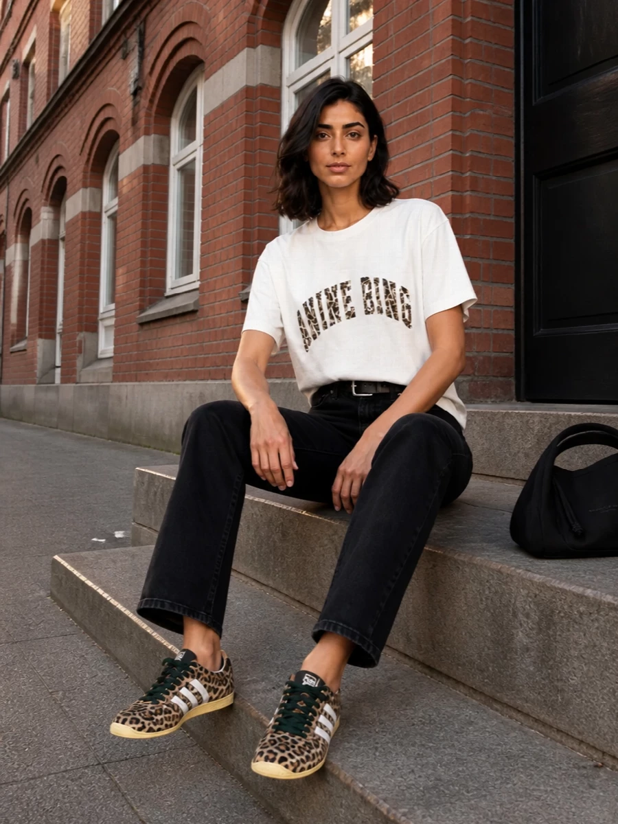 Anine Bing’s graphic tee is anchored by high-rise washed black denim for a clean, leg-lengthening base with a subtle rock attitude. Leopard sneakers echo the logo print without overpowering it, while a sculpted black shoulder bag and precise leather belt keep the whole outfit sharp and city ready. The result is relaxed streetwear with just enough structure for coffee runs that turn into late drinks.