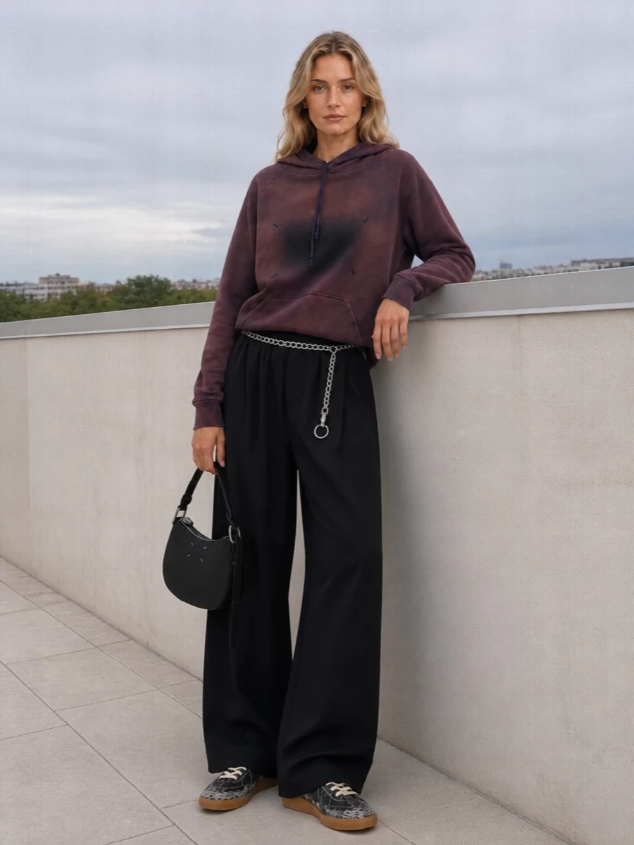 All about moody ease, this pairing leans into Margiela’s washed plum hoodie against deep black wide-leg trousers for a relaxed, elongating line. Crackled black sneakers keep the attitude soft but studied, while a compact crescent bag and silver chain belt give just enough structure and shine for studio days that stretch into late dinners.