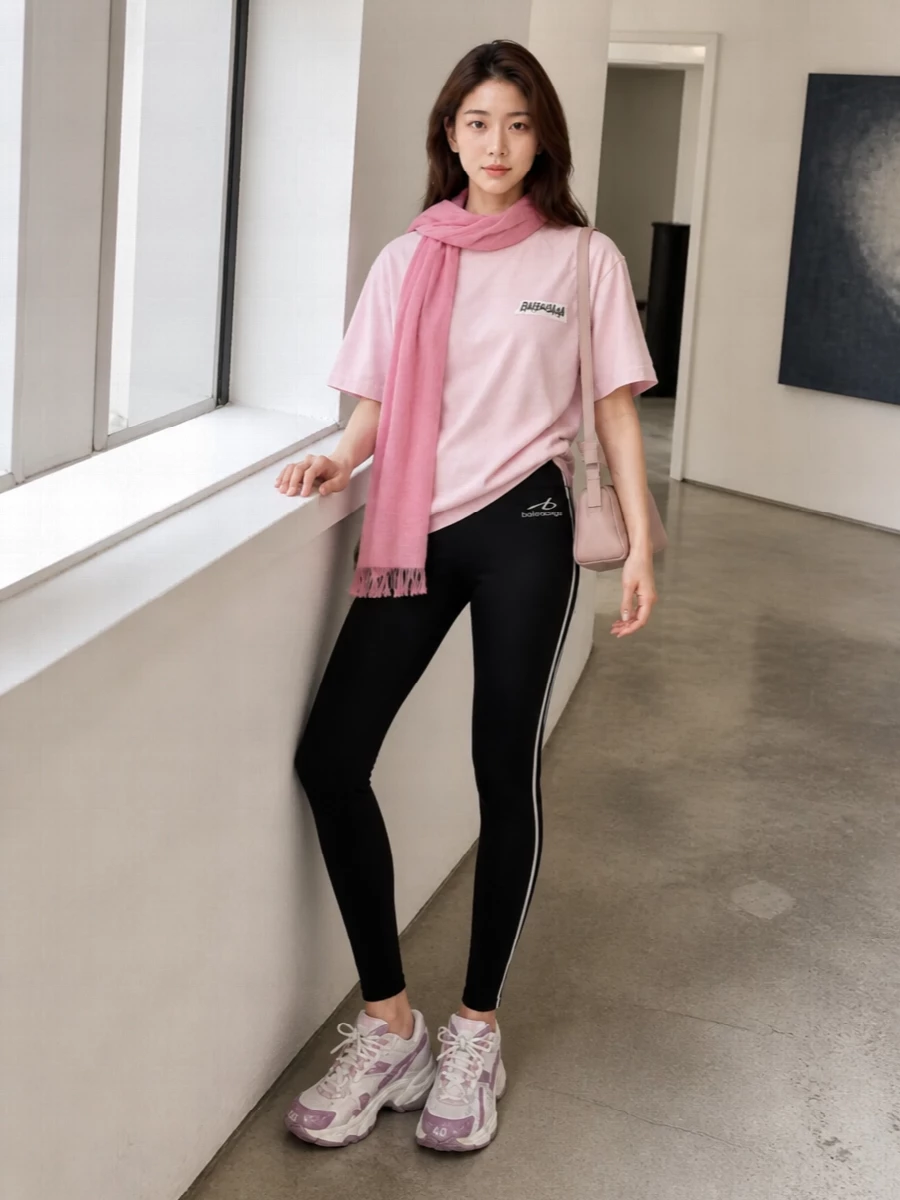 A blush Balenciaga tee sits over sleek black logo leggings for a clean, athletic base in that signature street-led proportion. Chunky white and lilac runners keep the mood technical yet playful, while a slim dusty-blush shoulder bag and rose-pink scarf echo the tee’s softness for a tightly edited pink‑and‑black city uniform.