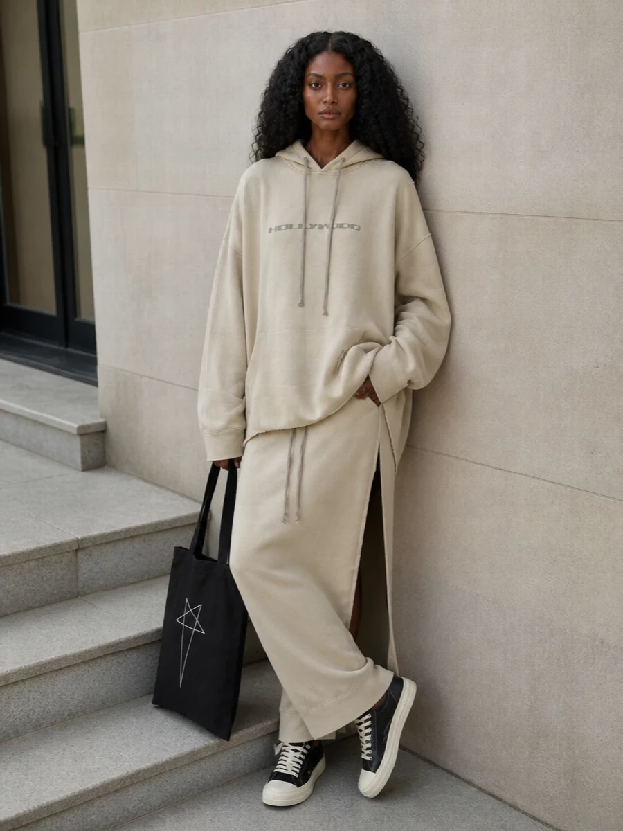A sun-bleached Rick Owens DRKSHDW hoodie falls over a cream column bottom, keeping everything long, pale and quietly graphic. Black and off-white sneakers echo the print and drawstrings, while a slim black tote adds a final, linear contrast so the volume reads intentional rather than sloppy. The result is slouched streetwear in soft neutrals with a precise, elongated silhouette.
