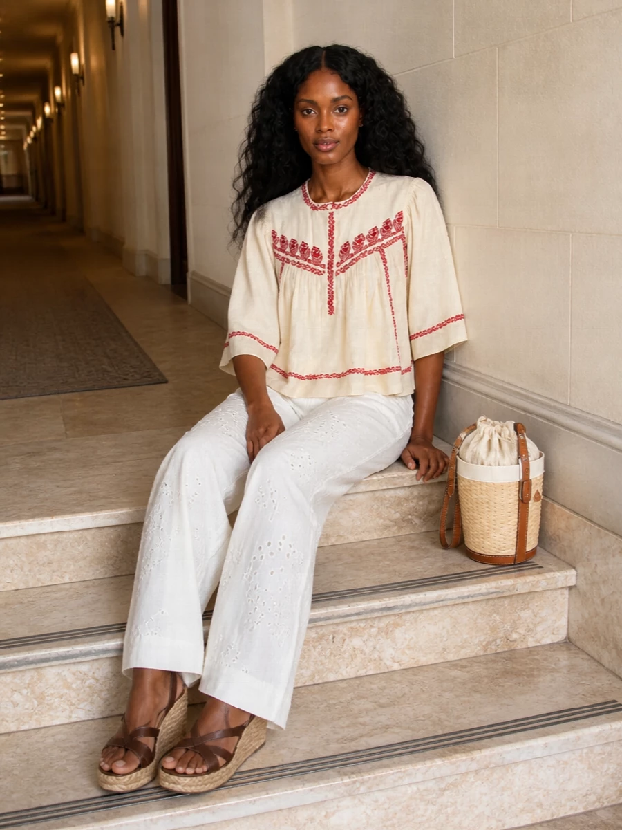 Romantic folk embroidery meets quiet-luxury ease with the blouse floating over wide-leg broderie trousers in soft ivory-on-ivory. Dark suede raffia wedges ground the softness, while a structured raffia mini bucket keeps the mood sunlit and polished. It is an easy summer outfit for terraces, garden parties and long city days that turn into dinner.