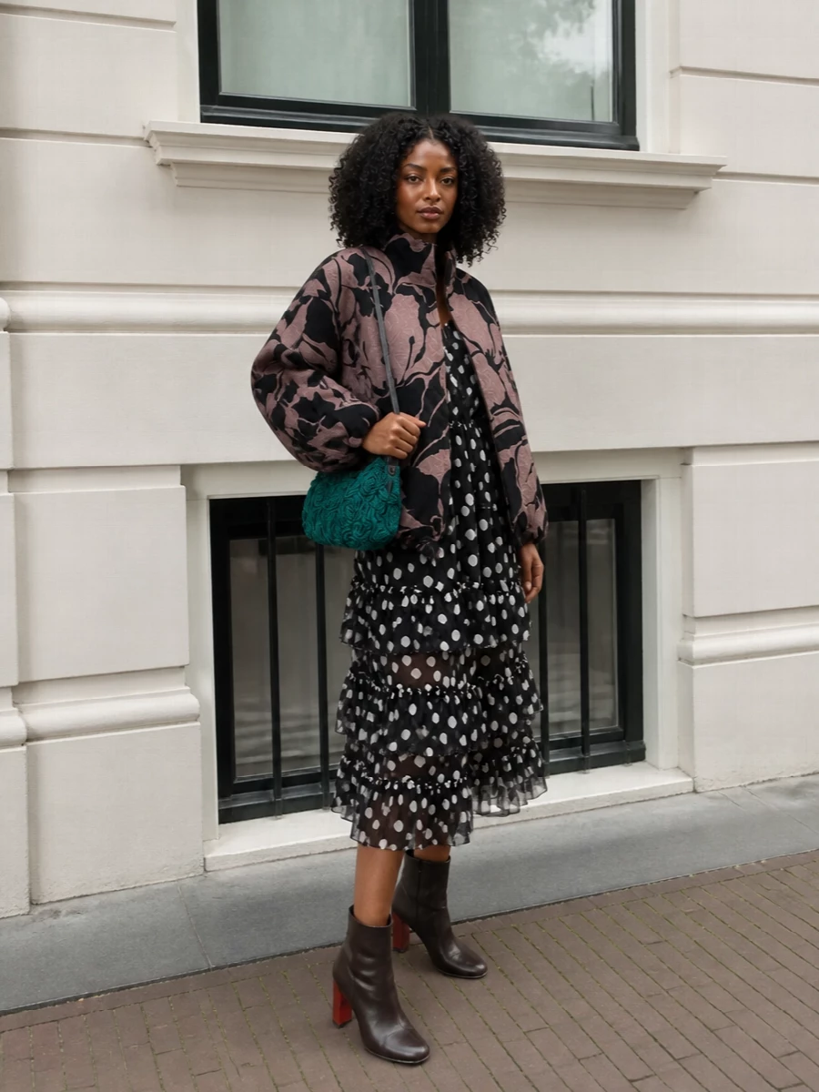 I am building around the coat with a dress rather than separates, so the volumes layer cleanly and the eye reads one continuous, graphic column under the cocoon. The polka-dot midi and sleek ankle boots keep everything in the same romantic, gallery-ready register, while the emerald rosette bag introduces a single saturated accent that still feels collected rather than loud. It is an art-world evening ensemble that lets the coat stay hero but finishes every angle with considered texture.