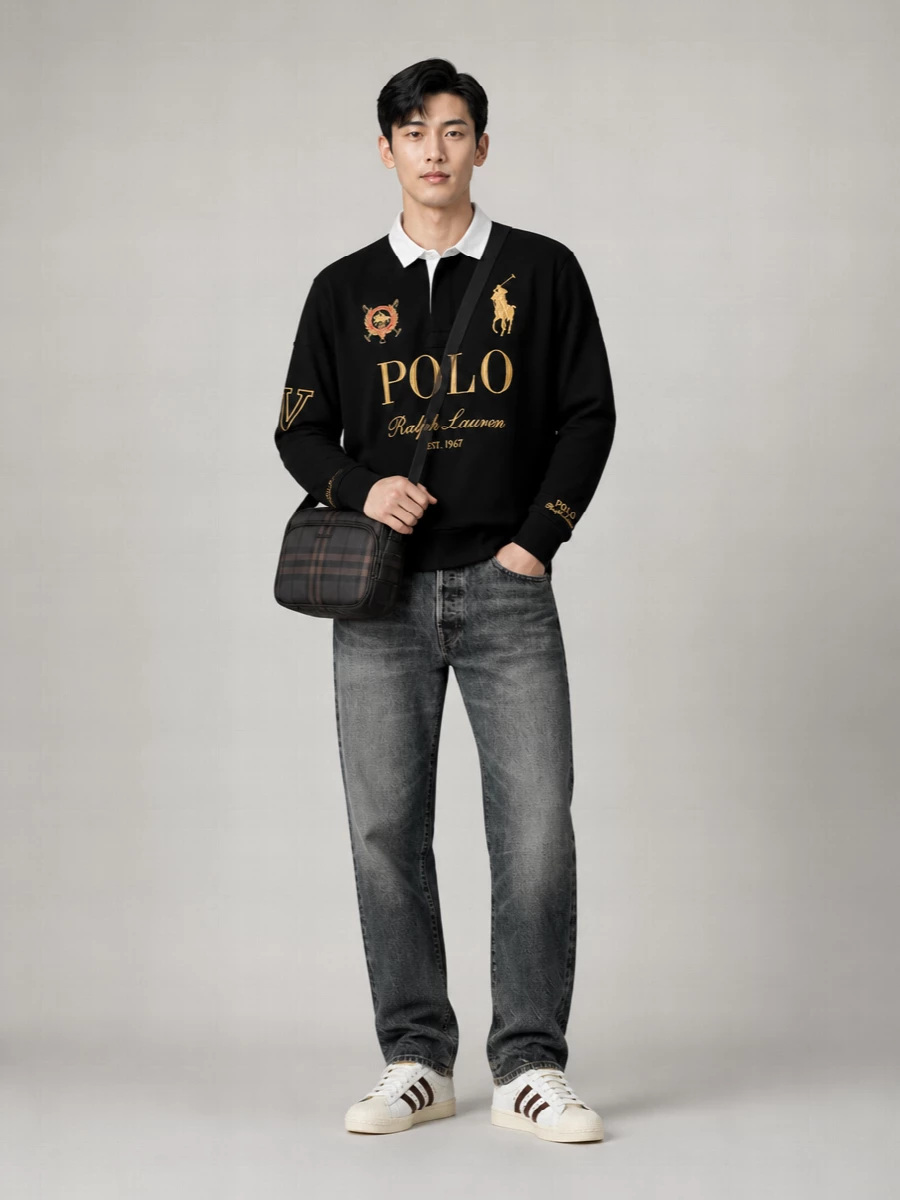 A graphic black rugby with gold cresting sits over faded charcoal denim for a sharp collegiate street mood. Textured shell-toe sneakers and a shadow-check crossbody keep the palette tight and premium, with the scarf echoing the team colors in a subtle hit of pattern.