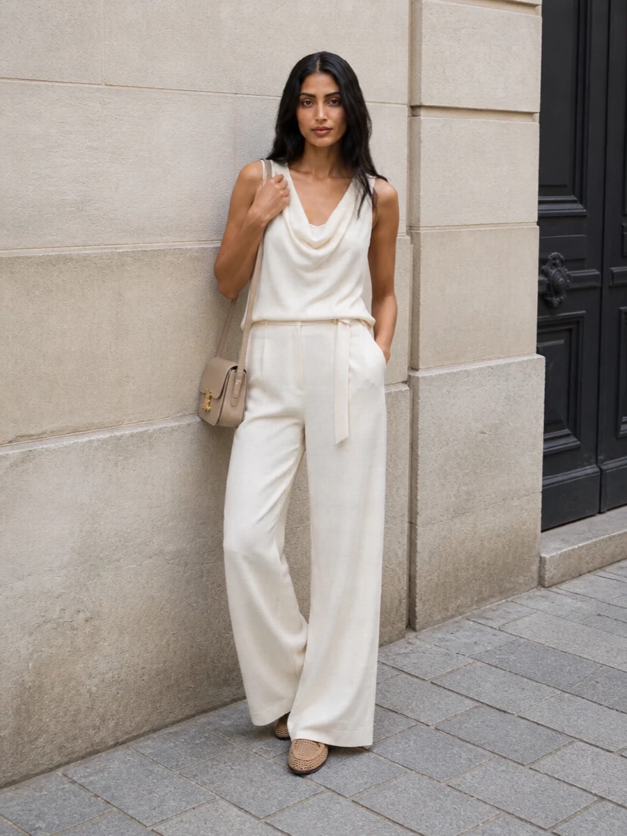 Tonal ivory wide-leg trousers are paired with a draped cowl-neck tank in the same soft hue for an elongated, fluid column that feels quietly luxurious yet effortless for SS. Sand perforated loafers introduce breathable structure at the base, while a small warm-beige Celine shoulder bag adds a refined, sculpted counterpoint that keeps the look polished from daytime meetings to dinner. The result is an understated, modern uniform built on light, harmonious neutrals and immaculate tailoring.