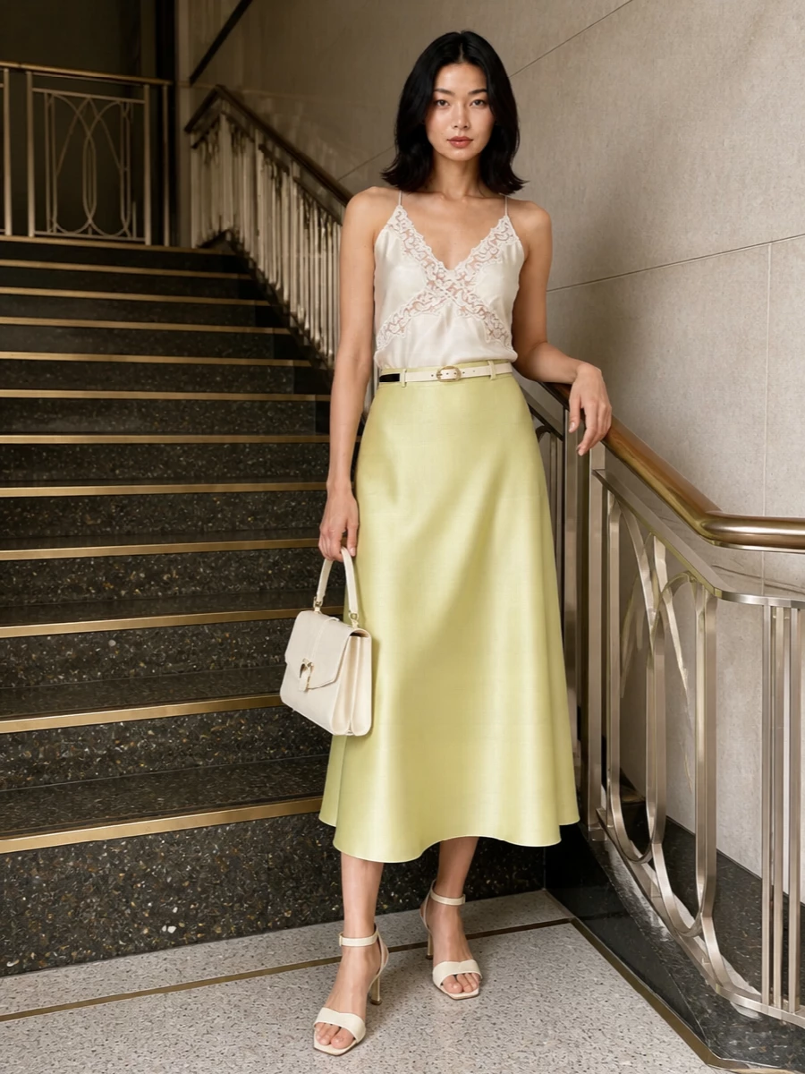 Pale lemon satin meets ivory in a quietly luminous pairing that feels romantic yet restrained for summer weddings or terrace dinners. The fluid camisole and high-waisted A-line skirt create a long, uninterrupted column, subtly defined by a slim belt. Cream sandals and an ivory top-handle bag keep the focus on light, liquid texture rather than hard contrast.