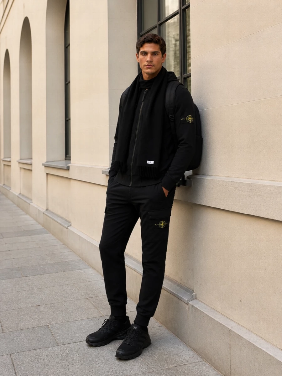 A monochrome Stone Island base that leans into clean technical streetwear, with the cargo jogger echoing the hoodie’s utility hardware and compass detailing. The black trail sneakers keep everything grounded and agile, while the nylon backpack and deep black scarf add just enough structure and texture to feel considered for city days and travel.