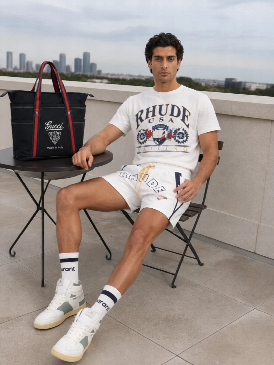 Rhude’s sailing tee sits on matching white logo shorts for a tight, regatta-ready story that feels graphic but easy. Sage-toned court sneakers and a navy Gucci tote keep things grounded in upscale sport, with striped socks adding a subtle collegiate hit. It is a clean, sun-forward uniform for city days that can slide straight to the marina or pool deck.