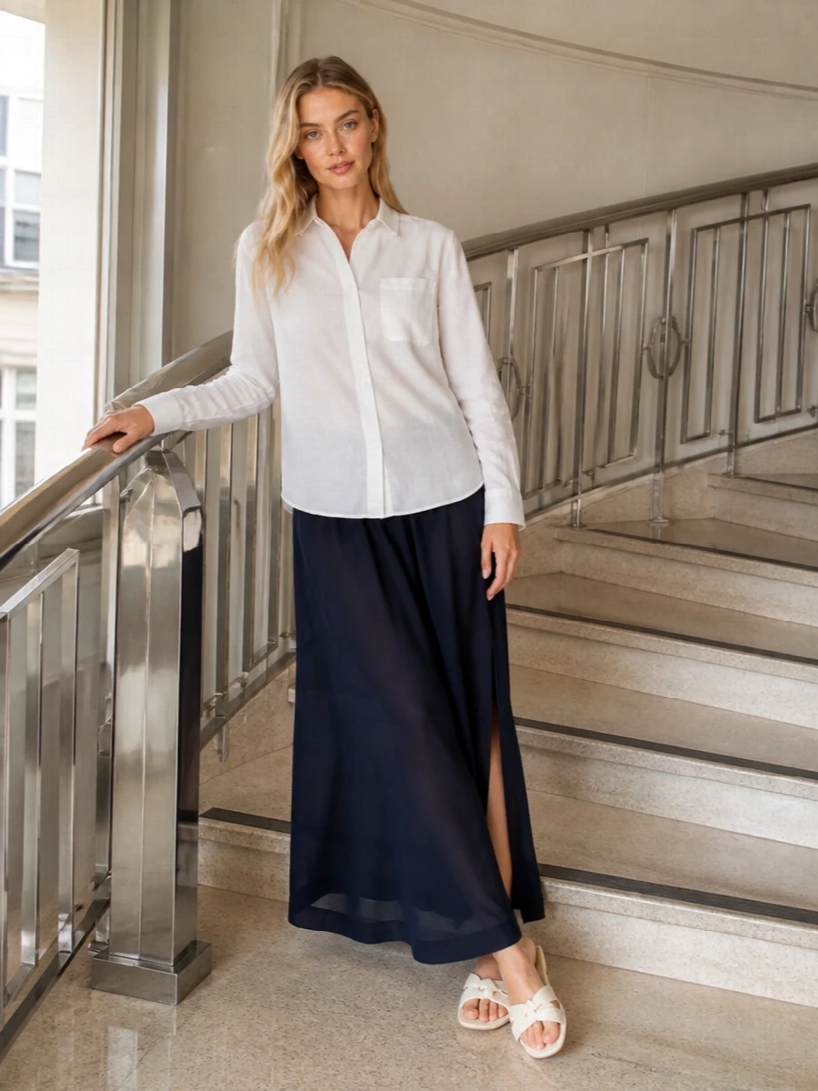 A sheer navy maxi grounded with a crisp white shirt keeps the mood clean and coastal, ideal for moving between beach and terrace. Ivory slides and a structured raffia tote keep the palette light while maintaining that understated, quietly polished feel. The proportions stay relaxed and long, so the whole outfit feels breezy without losing structure at the waist and collar.