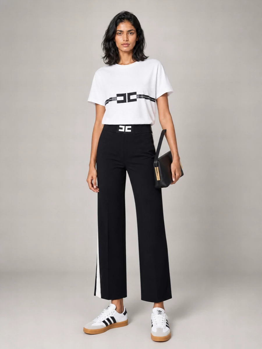 A graphic black and white base keeps this tee front and center, anchored by logo-waist knit trousers that nod to track styling without losing structure. Clean terrace sneakers and a sharp black shoulder bag keep the mood sporty yet refined, while a slim gold-buckle belt adds a hint of Italian polish without breaking the minimal line. It is an easy city uniform that reads relaxed, branded and quietly precise all at once.
