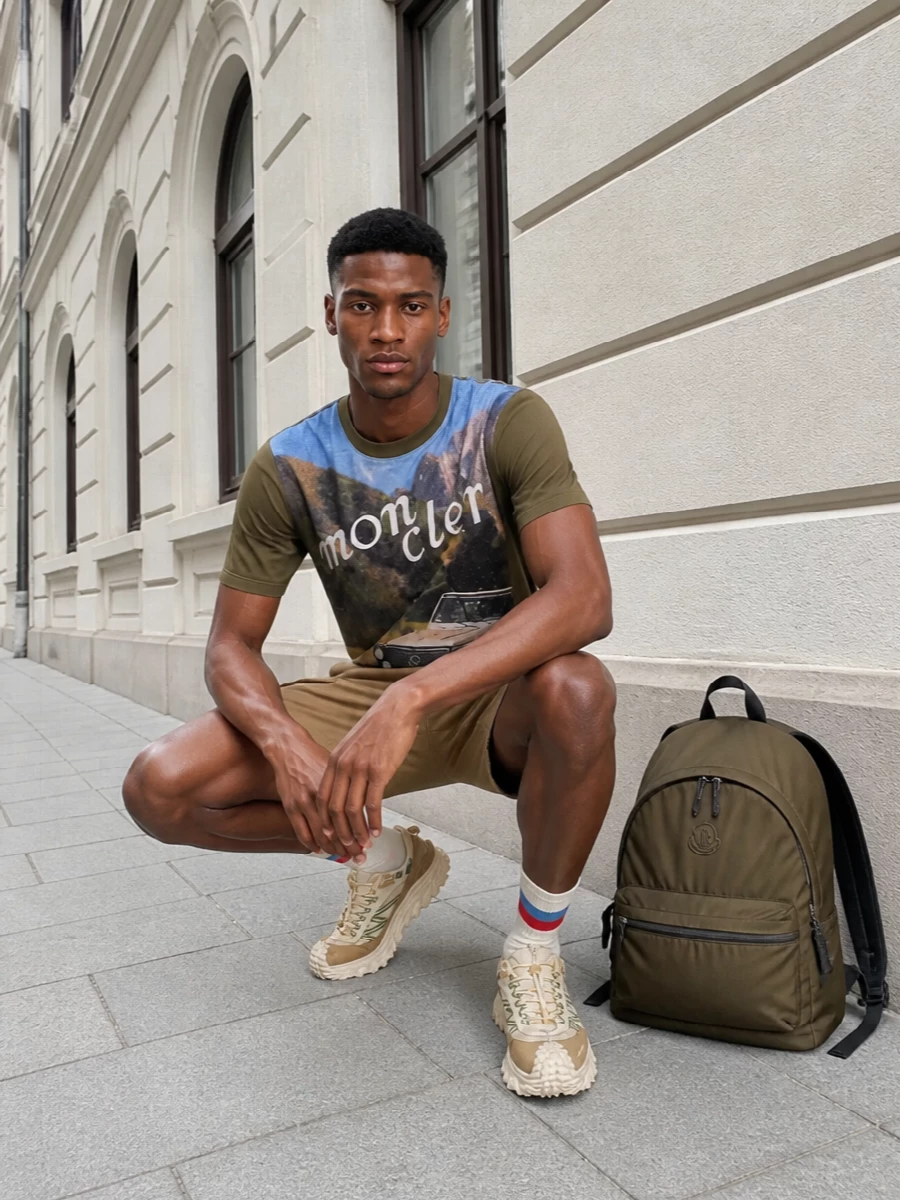 An easy alpine-inspired outfit that leans into the tee’s vintage road-trip energy with camel cargo shorts and trail-ready sneakers in cream, sand and olive. The technical fabrics, elastic waists and lugged sole keep it practical for real movement, while the earthy palette stays tightly edited around greens, tans and sky tones. A khaki Moncler backpack and striped socks complete a polished, outdoorsy streetwear set that feels ready for city hikes and weekend escapes alike.